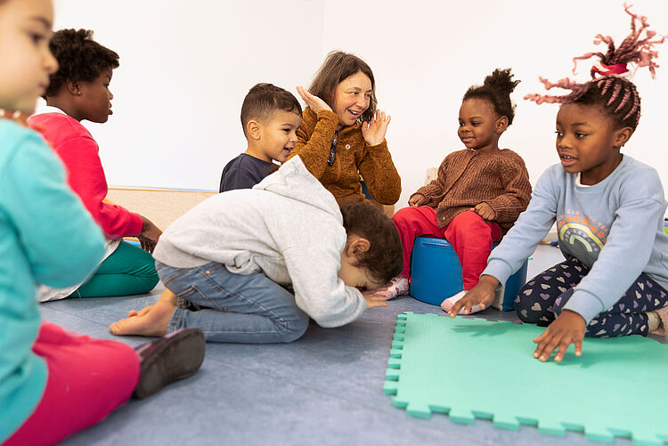 Kindertagesstätte Eine Erzieherin sitzt und spielt mit mehreren Kindern auf dem Boden. Die Kinder lachen.