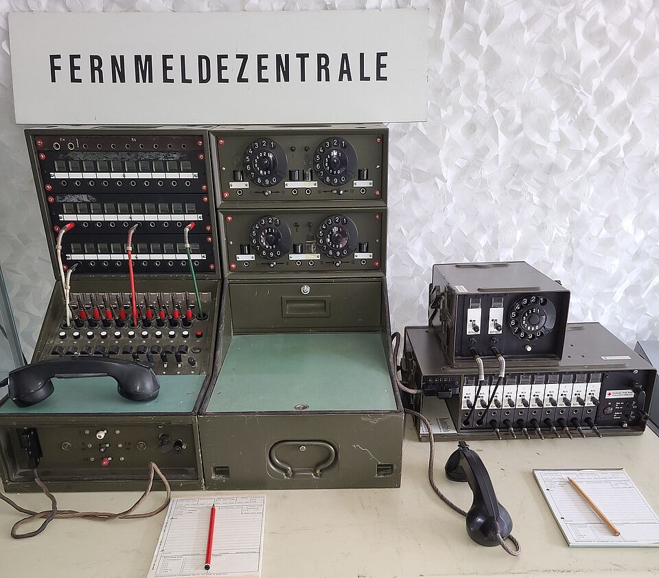 Historische Funk- und Fernmeldegeräte auf einem Tisch mit einem Schild „Einsatzführung“, einem geöffneten Einsatzbuch und Einsatzunterlagen.
