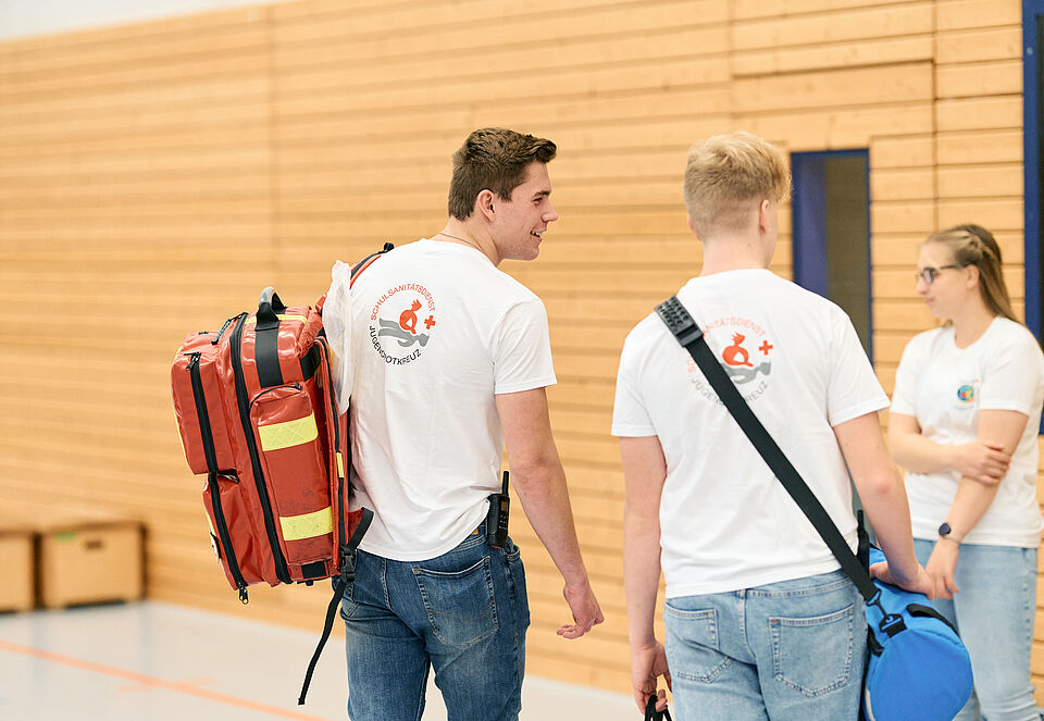 Drei Schulsanitäter stehen mit einem Notfallrucksack in einer Sporthalle.