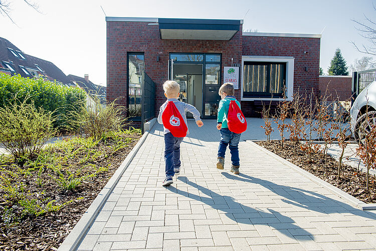 Zwei Kinder mit roten DRK-Turnbeuteln auf dem Rücken laufen zum Eingang der Kindertagesstätte.