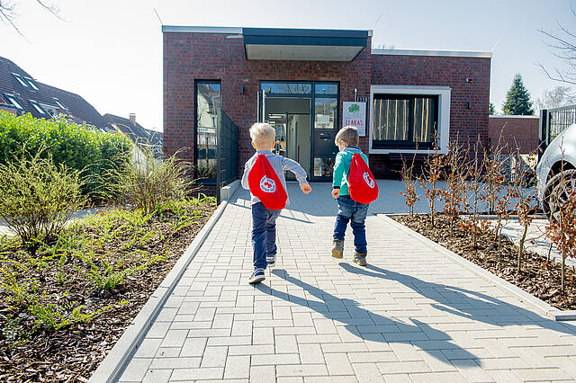 Zwei Kinder mit roten DRK-Turnbeuteln laufen zum Eingang der Kindertagesstätte.