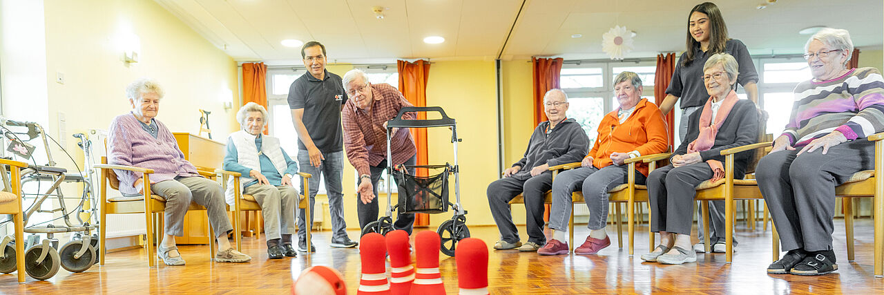 Kegeln in der Cafeteria Mehrere Senior*innen sitzen im Halbkreis zusammen. In der Mitte spielt ein Sozialarbeiter mit einem Bewohner Kegeln.