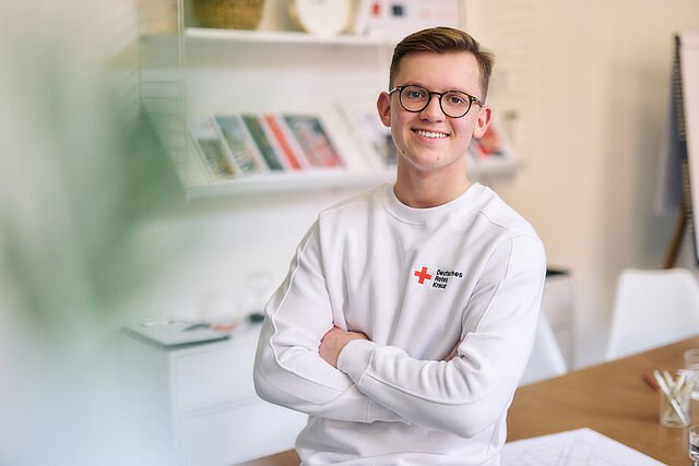 Ein junger Mitarbeiter des DRK trägt einen weißen Pulli mit DRK-Logo und lehnt mit verschränkten Armen an einem weißen Tisch.