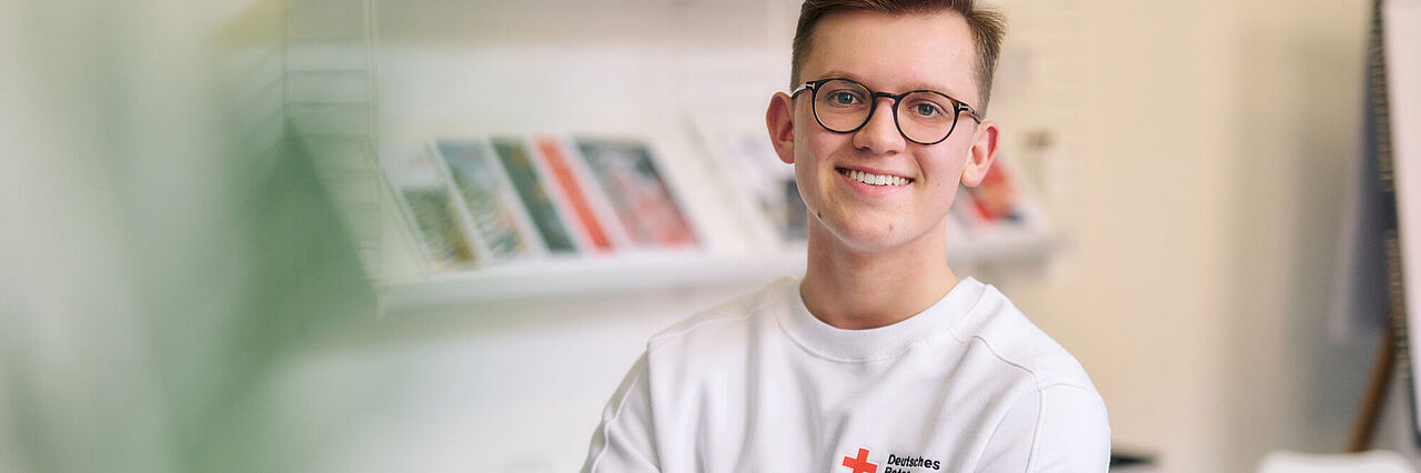 Ein junger Mitarbeiter des DRK trägt einen weißen Pulli mit DRK-Logo und lehnt mit verschränkten Armen an einem weißen Tisch