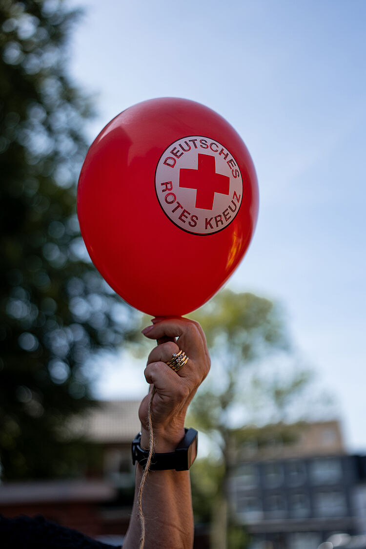 Person hält roten Luftballon mit DRK-Logo in die Luft. Am Handgelenk ist eine DRK-Watch.