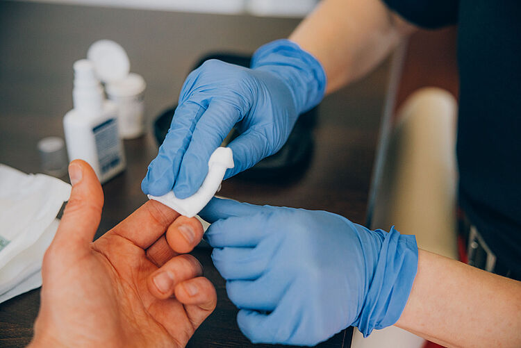 Hände in blauen medizinischen Handschuhen versorgen eine andere Hand mit einem Tupfer.