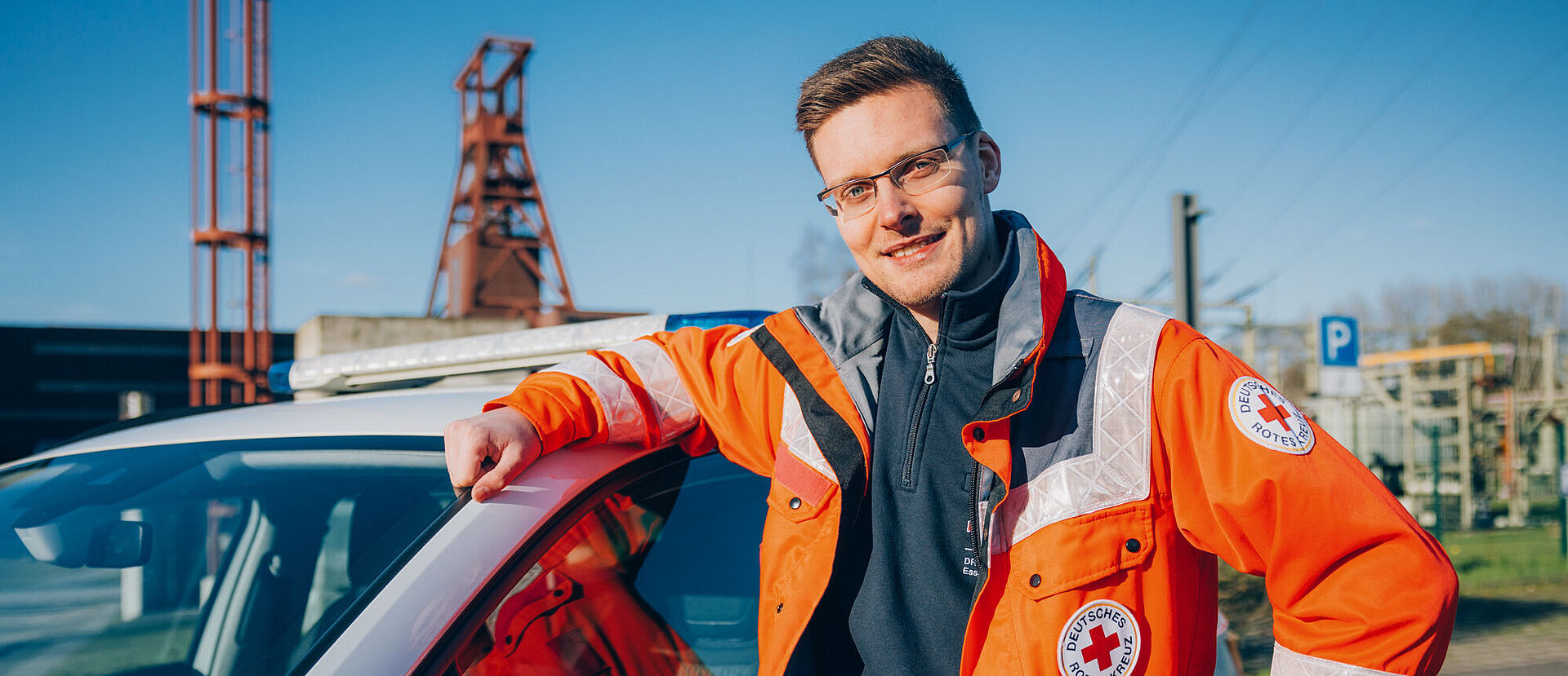 Ein lächelnder junger Mitarbeiter des DRK Essen lehnt in Einsatzkleidung an einem Auto des DRK. Im Hintergrund ist die Zeche Zollverein erkennbar.