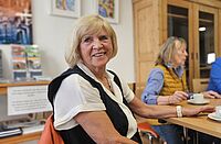 Blonde ältere Frau lächelt in die Kamera. Sie sitzt mit zwei weiteren Frauen an einem großen Holztisch auf dem eine rote Kaffeekanne steht.