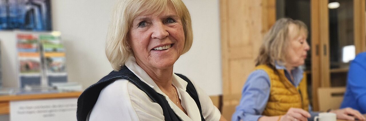 Zentrum 60plus Blonde ältere Frau lächelt in die Kamera. Sie sitzt mit zwei weiteren Frauen an einem großen Holztisch auf dem eine rote Kaffeekanne steht.