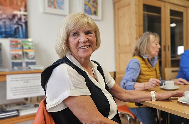 Blonde ältere Frau lächelt in die Kamera. Sie sitzt mit zwei weiteren Frauen an einem großen Holztisch auf dem eine rote Kaffeekanne steht.