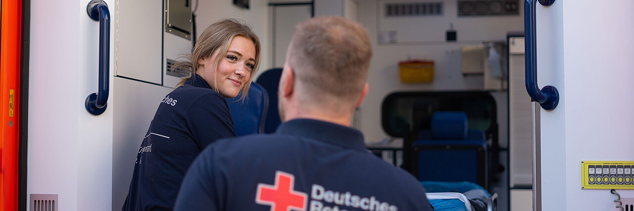 Zwei Mitarbeitende, eine Frau und ein Mann, steigen in einen Rettungswagen ein. Blick durch die große Tür ins Innere des Wagens.