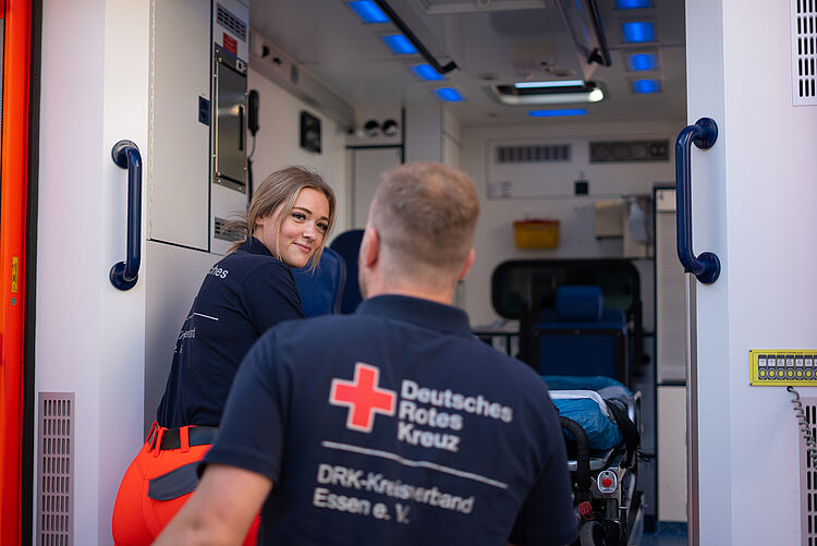 Zwei Mitarbeitende, eine Frau und ein Mann, steigen in einen Rettungswagen ein. Blick durch die große Tür ins Innere des Wagens.