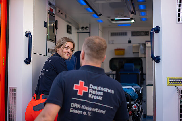 Zwei Mitarbeitende, eine Frau und ein Mann, steigen in einen Rettungswagen ein. Blick durch die große Tür ins Innere des Wagens.