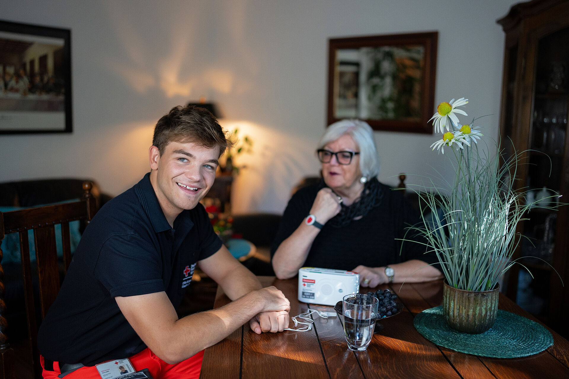 DRK Essen Hausnotruf Eine Rettungskraft und eine Seniorin sitzen an einem Tisch. Die Rettungskraft lächelt in die Kamera. Auf dem Tisch steht die Basisstation des Hausnotrufs; die Seniorin trägt den Funksender.
