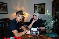 Eine Rettungskraft und eine Seniorin sitzen an einem Tisch. Die Rettungskraft lächelt in die Kamera. Auf dem Tisch steht die Basisstation des Hausnotrufs; die Seniorin trägt den Funksender.